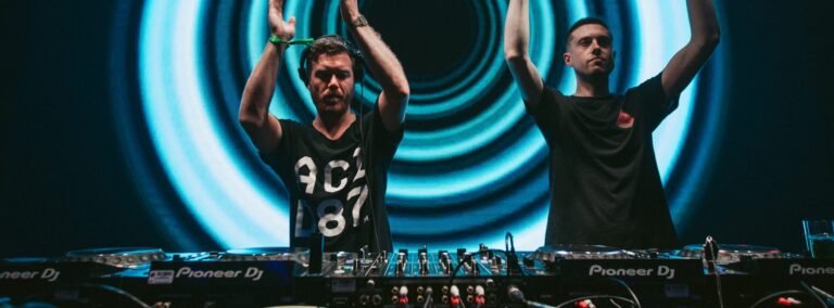 Two djs raising their hands in excitement while performing at a concert, surrounded by dj equipment and intense stage lighting.