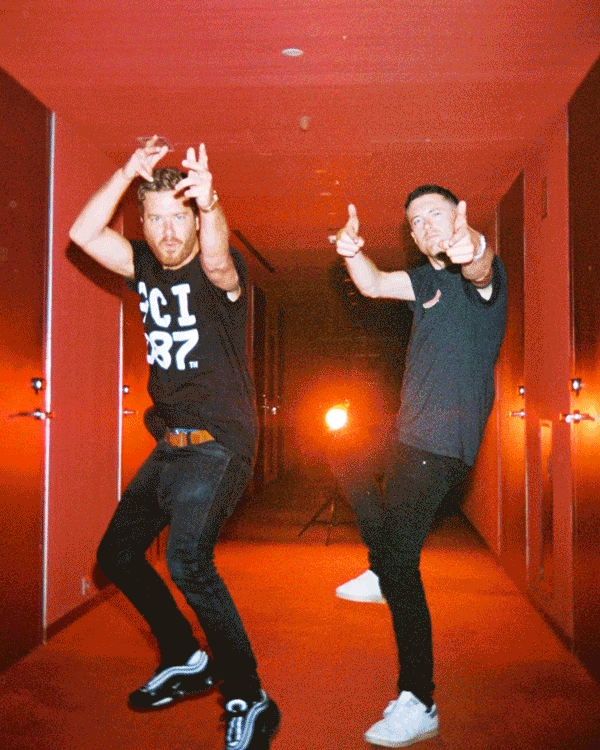 Two men dancing playfully in a red-lit corridor, making finger guns and playful gestures towards the camera.