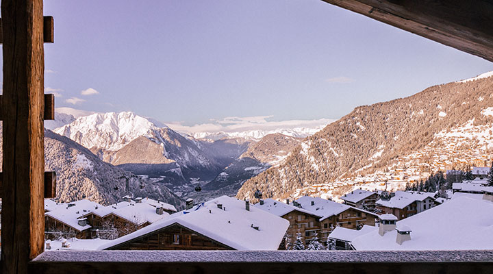 Vista desde un balcón de madera sobre tejados nevados y laderas de montaña, con un cielo despejado y picos iluminados por el sol al fondo.