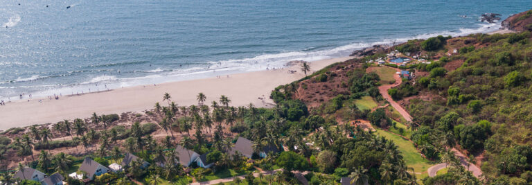 Aerial view of a tropical beach with white sand, scattered palm trees, green vegetation, and small buildings near the shoreline; people walk along the edge of the calm sea.