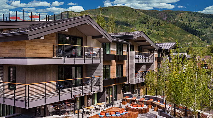 Moderno albergue de montaña con exterior de madera, grandes balcones y zona de estar al aire libre. Rodeado de verdes árboles y colinas bajo un cielo azul con nubes.
