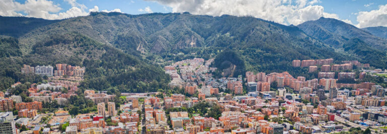 A panoramic view of a city with numerous colorful buildings at the base of lush, green, mountainous hills under a partially cloudy sky. The urban area is densely packed, contrasting with the natural landscape of the hills behind.