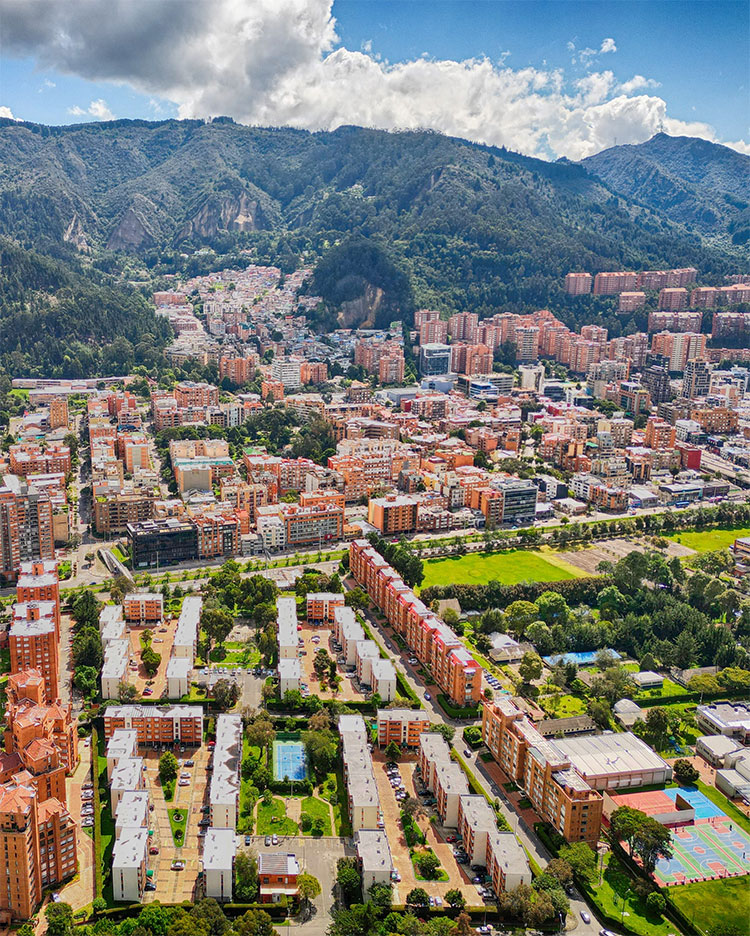 Aerial view of a cityscape featuring dense residential neighborhoods with multistory buildings, green parks, and tree-lined streets. In the background, there are forested mountains under a partly cloudy sky. The area looks vibrant and well-planned.