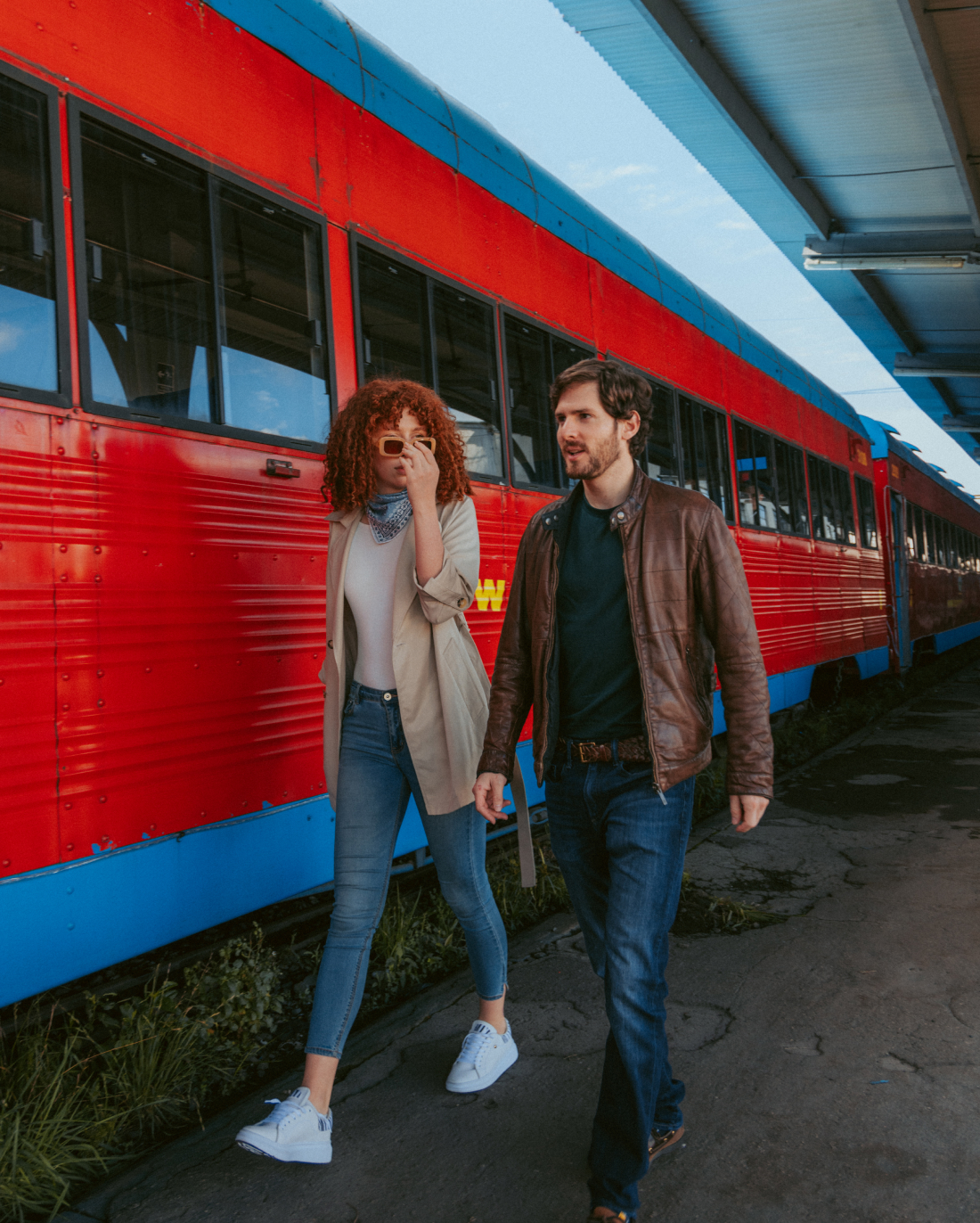 A woman with curly hair and sunglasses and a man with a beard walk along a train platform beside a bright red and blue train. She wears a beige coat and jeans, while he wears a brown leather jacket and jeans. The sky is visible through the platform roof.