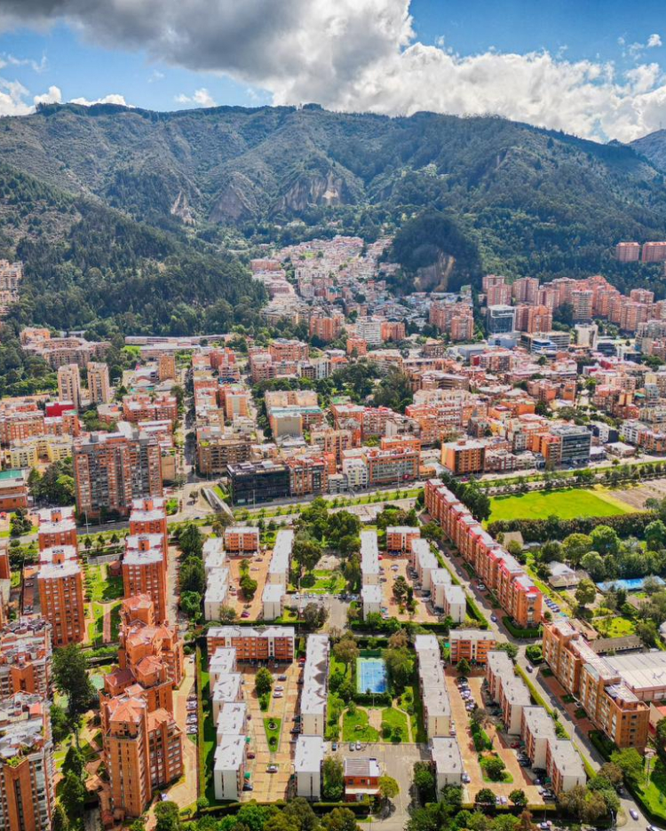 Aerial view of a city with a mix of colorful residential buildings, green spaces, and tree-lined streets. The city is surrounded by lush, hilly terrain under a partly cloudy sky, creating a scenic backdrop.