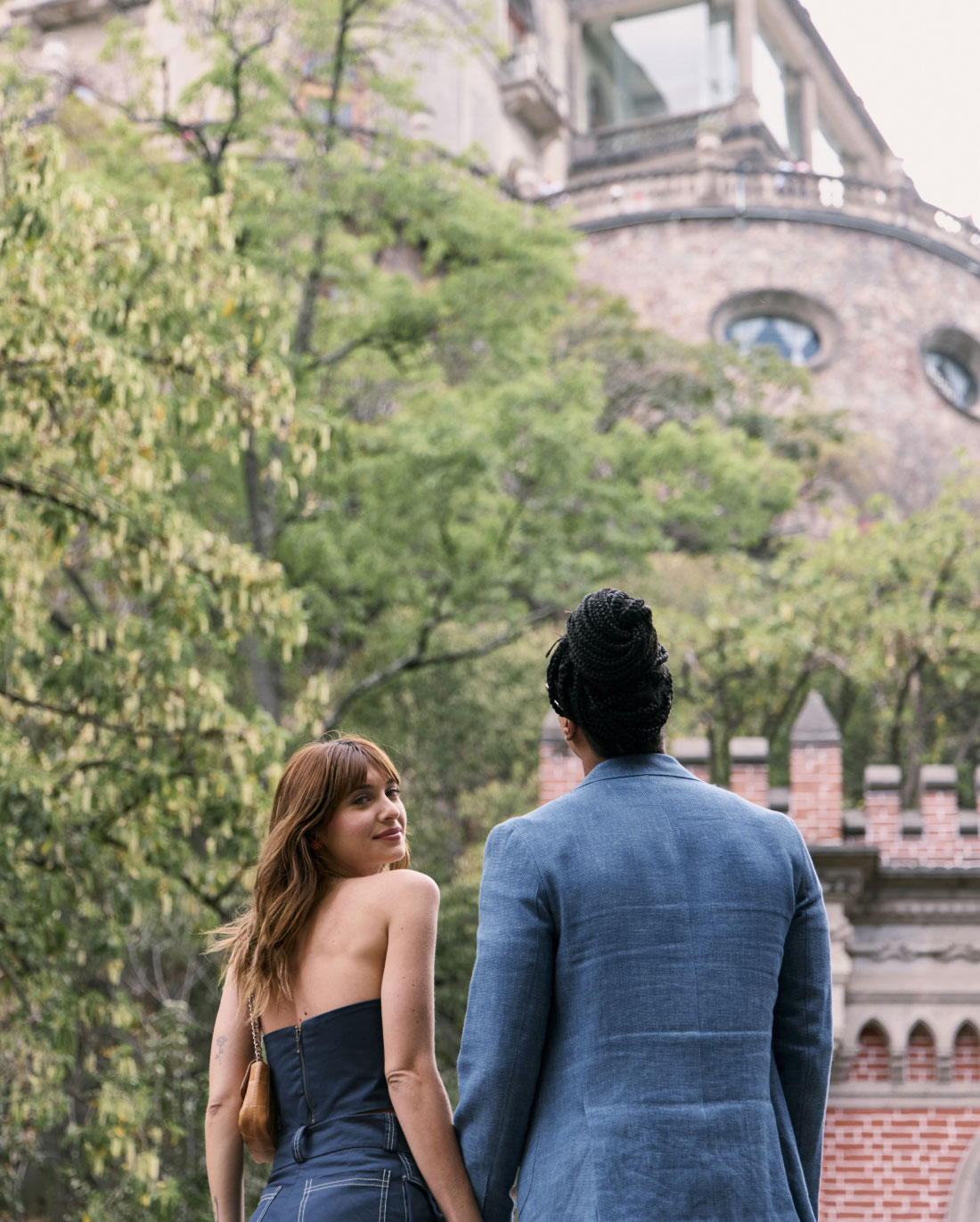 A woman and a man, both dressed in formal attire, stand closely together, holding hands and looking upwards. They are outdoors, with leafy trees and a historic-looking brick building with ornate architecture in the background.