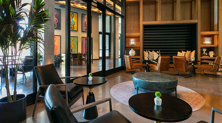 A modern lounge area featuring black leather chairs, round black tables with small plants, and a circular ottoman. In the background are wall art, large windows, and a cozy seating nook with cushions. The space is bright, with hardwood panels and polished floors.