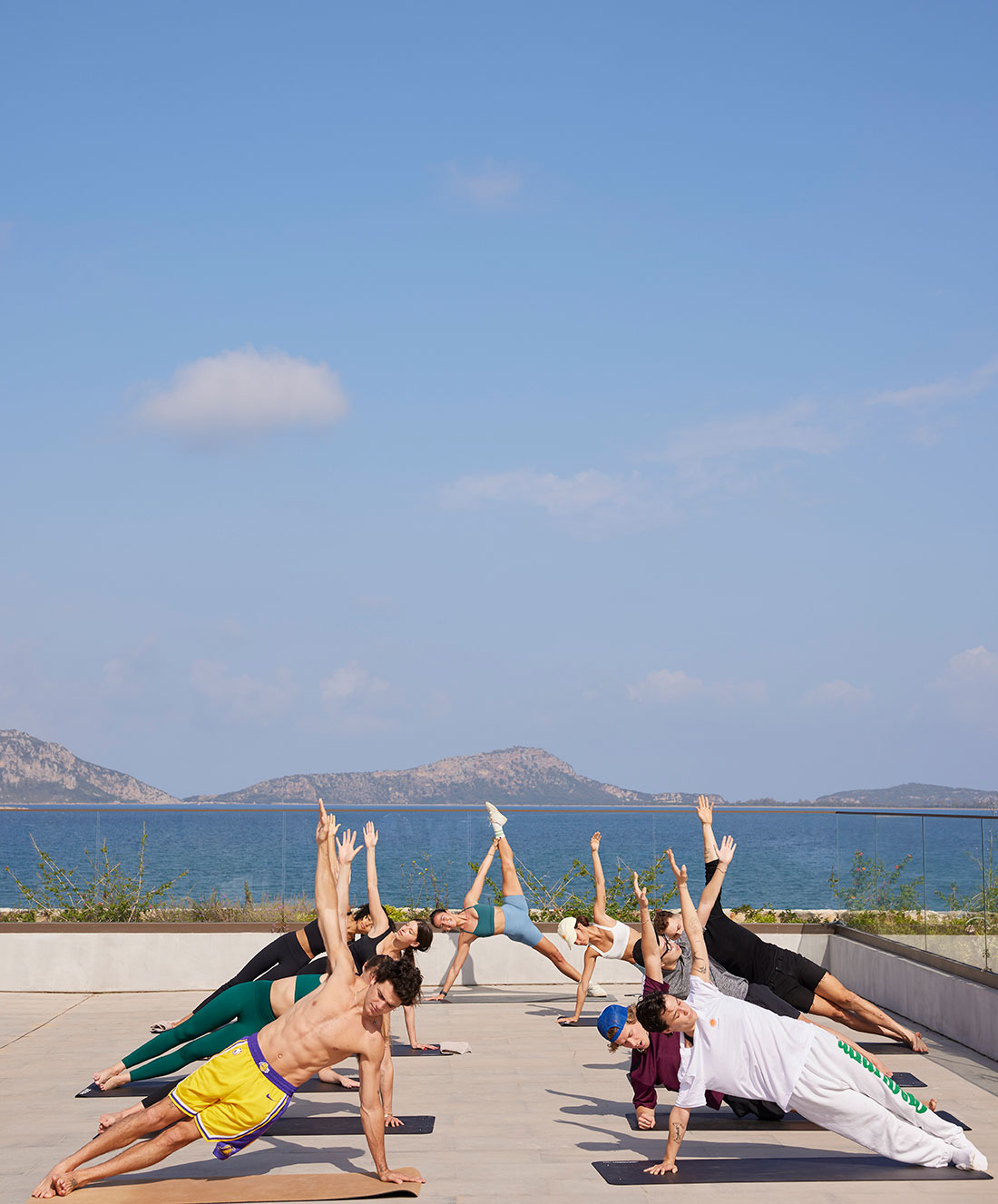 Un groupe de personnes pratique le yoga en plein air par une journée ensoleillée, effectuant des poses de planche sur le côté sur une surface carrelée avec la mer, les montagnes lointaines et le ciel bleu en arrière-plan.