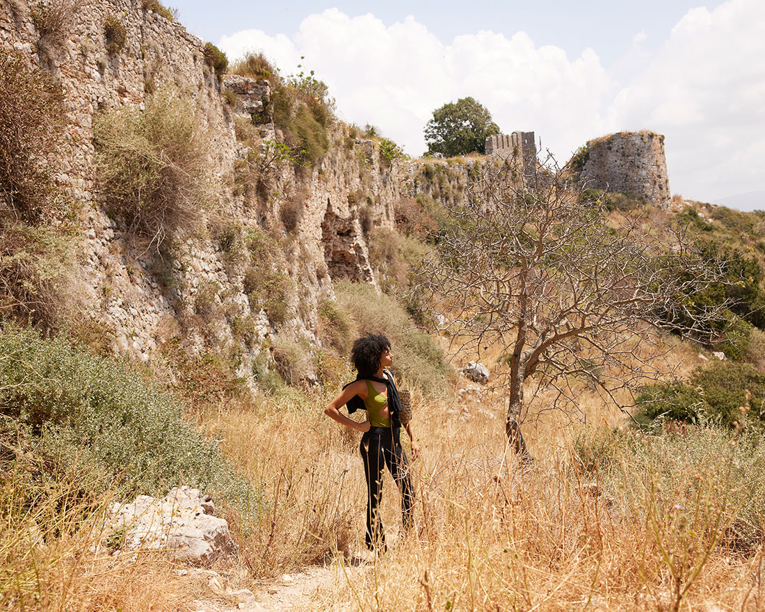 Une personne aux cheveux bouclés se tient dans l'herbe sèche, regardant les ruines d'un vieux mur de pierre sous un ciel lumineux, entouré d'une végétation sauvage et d'un arbre sans feuilles.