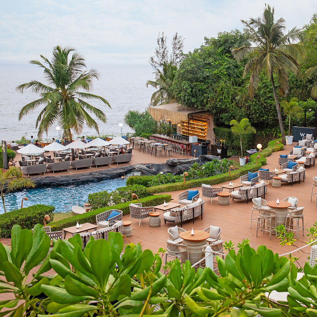 An outdoor restaurant and bar area with tables and chairs overlooks a pool and the ocean, surrounded by lush greenery and palm trees under a cloudy sky.