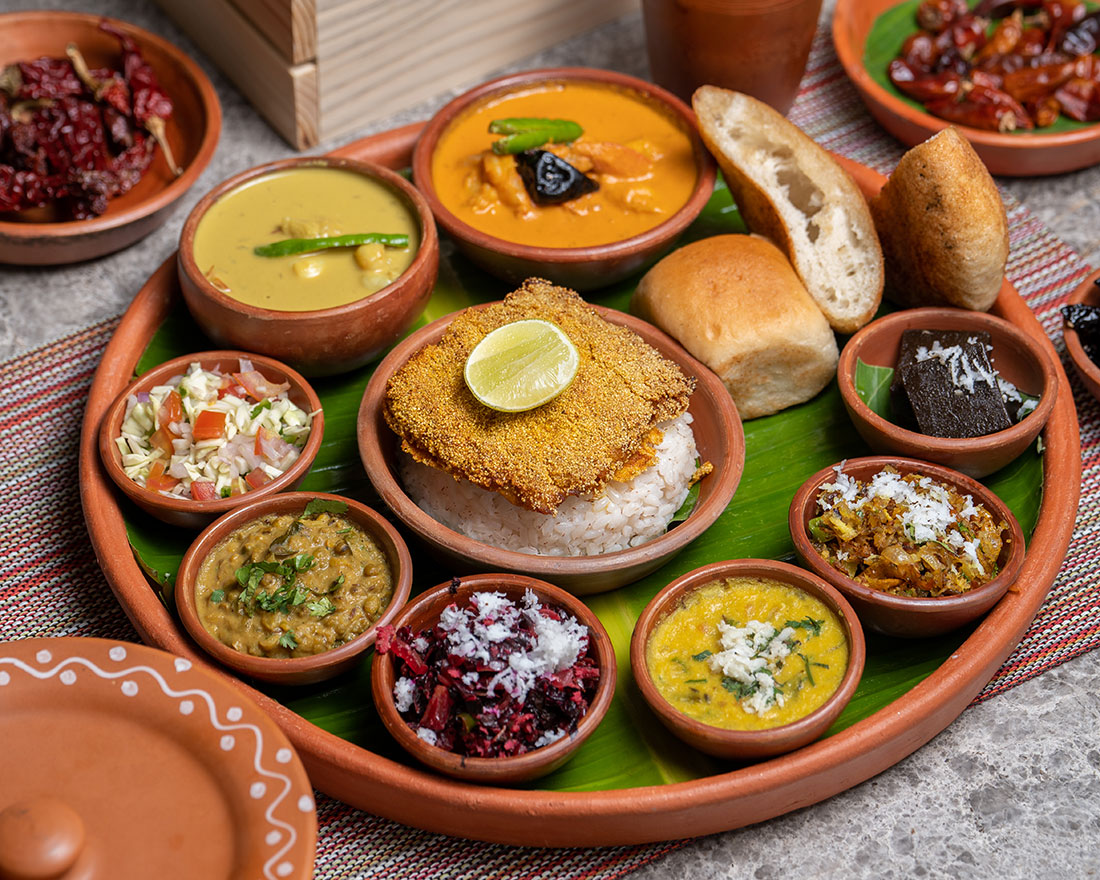 A traditional Indian thali served on a large plate with various bowls of curries, vegetables, lentils, rice topped with fried fish and a lemon slice, bread rolls, salad, sweets, and pickles, all arranged neatly on a banana leaf.