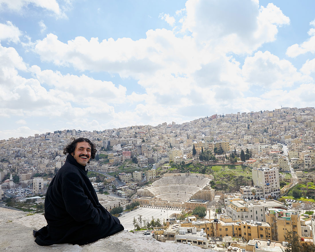 A person with dark hair and a mustache, wearing a black coat, sits on a ledge overlooking a city with densely packed buildings and a large ancient amphitheater under a partly cloudy sky.