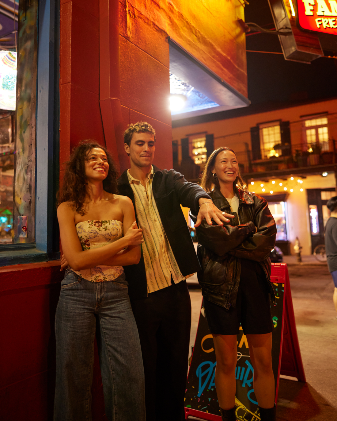 Three friends enjoying an evening out, standing and laughing together near a colorful street corner illuminated by neon lights.
