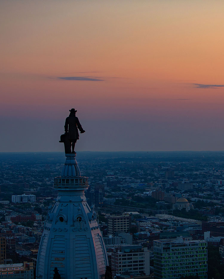 A sunset view over Philadelphia, featuring the silhouette of the William Penn statue atop the dome of City Hall, with the cityscape spread out below under a colorful sky.