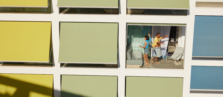 A man and a woman stand talking on a balcony of a modern building with pastel green and yellow window shades. The woman holds a drink, and the man points outward, both enjoying the outdoor space.
