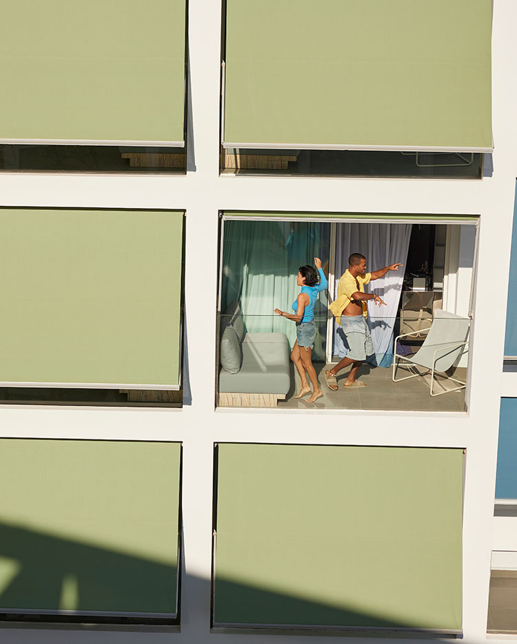 Un homme et une femme dansent ensemble sur un balcon, vu à travers une grande fenêtre d'un bâtiment dont les stores vert clair couvrent la plupart des autres fenêtres.