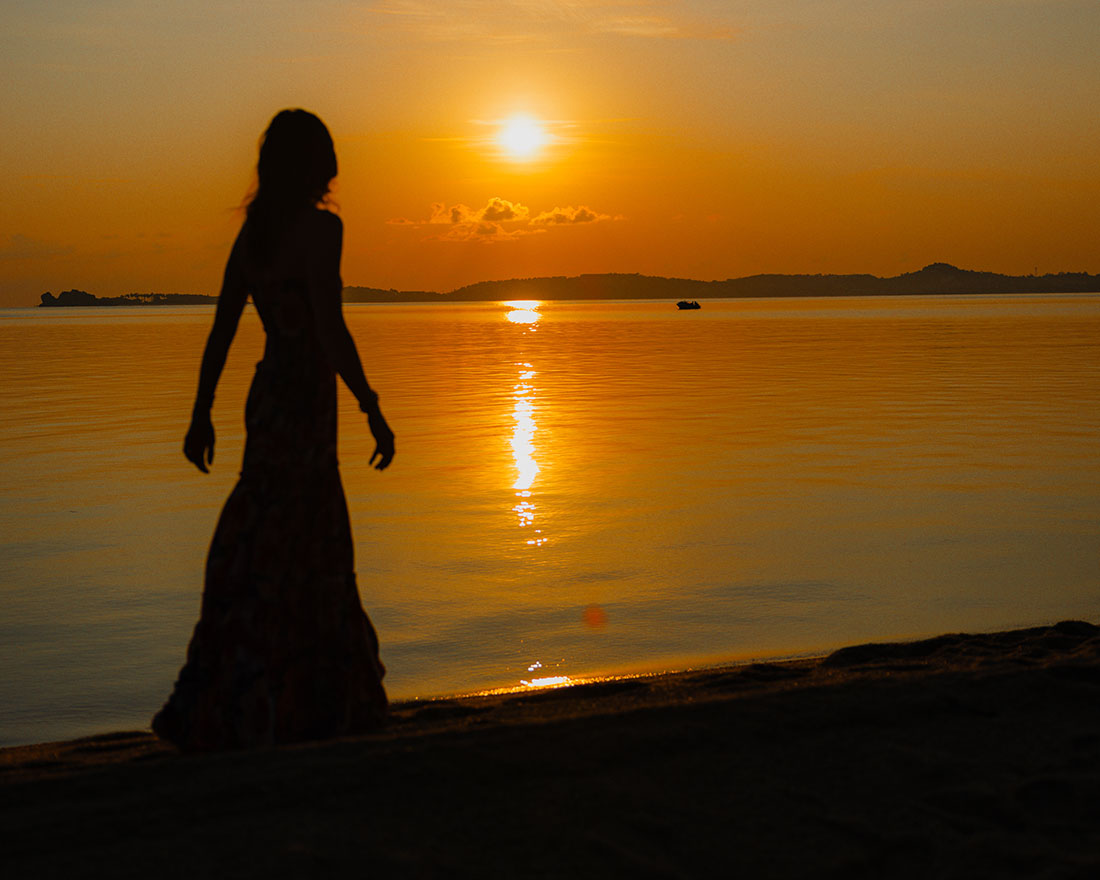 夕暮れの浜辺を歩くロングドレス姿の人物のシルエット。空には太陽が低く沈んでおり、その反射が穏やかな水面に揺らめいている。.