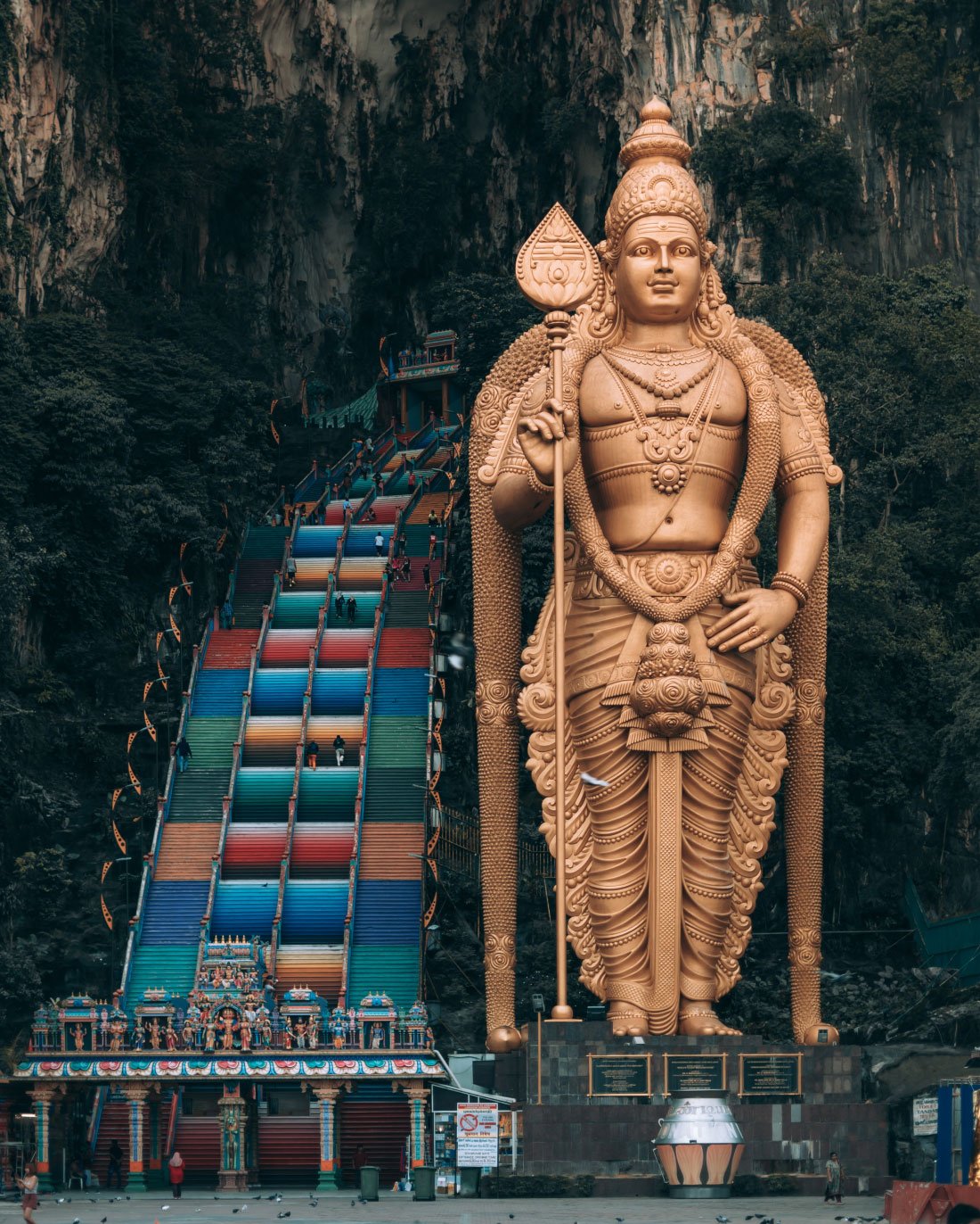 A towering golden statue of Lord Murugan stands majestically in front of a vibrant, multi-colored staircase leading to the Batu Caves. The statue is intricately detailed, and the lush, rocky backdrop of the caves adds to the grandeur of the scene.
