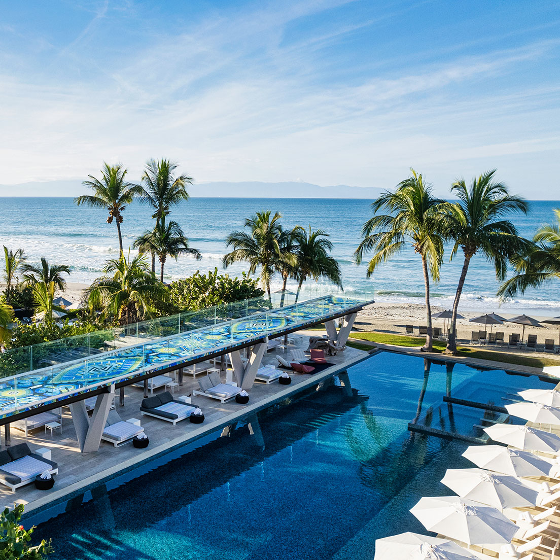 A luxurious beachfront resort features an expansive pool with sunbeds and umbrellas, surrounded by tall palm trees. The pool overlooks a serene ocean, offering clear views of the blue sky and distant mountains.