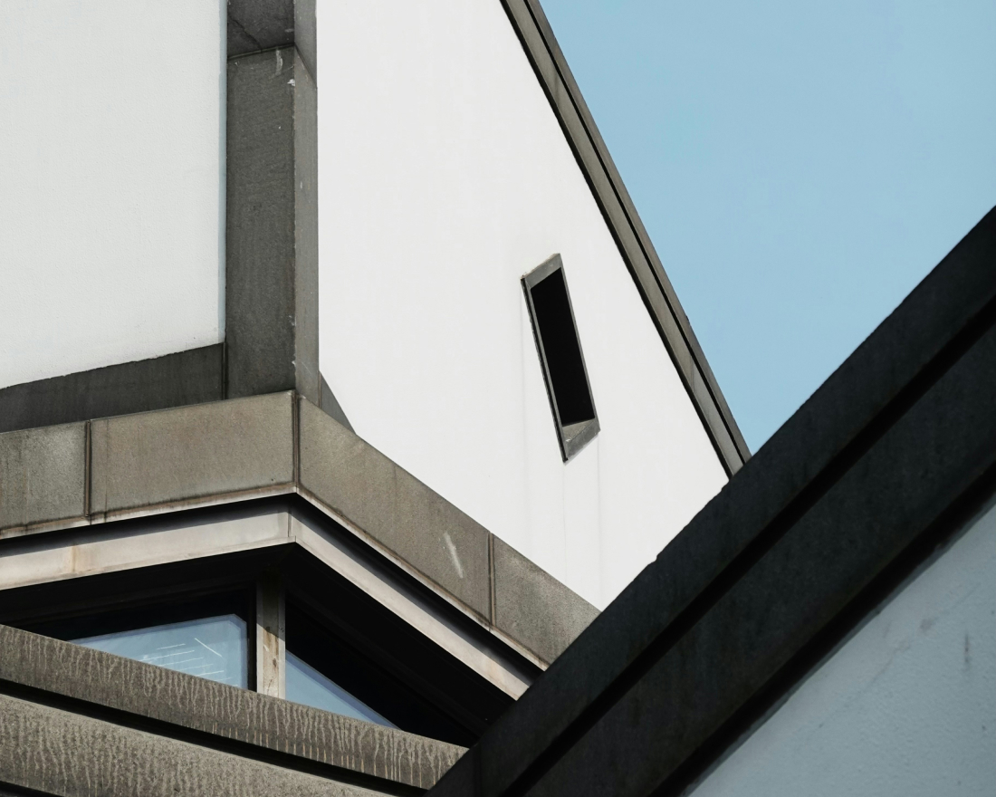 Ein minimalistisch anmutendes Gebäude mit eckigen Linien und Betonoberflächen ist vor einem klaren blauen Himmel zu sehen. Auf der weißen Fassade ist ein kleines rechteckiges Fenster zu sehen, während andere Abschnitte dunklere, schattige Bereiche zur Schau stellen.