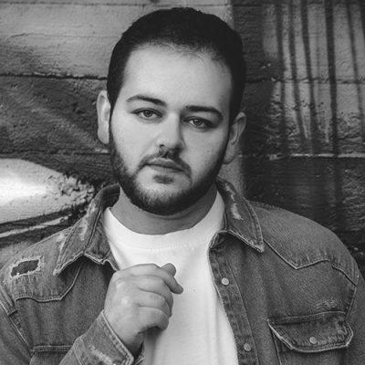 Black and white portrait of a young man with a beard, wearing a denim jacket over a white shirt, standing against a textured brick wall. he is looking directly at the camera.