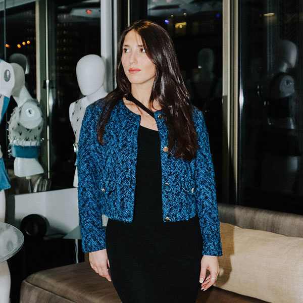 Woman with long dark hair wearing a black dress and blue jacket stands indoors. Mannequins with various outfits are visible in the background.