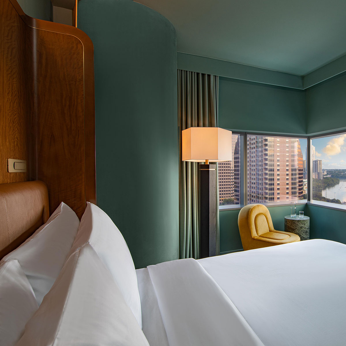 A luxurious hotel room features a neatly made bed with white linens, a yellow chair by the window, and a view of a city skyline and river. The walls are painted green, and a tall lamp stands beside the window.