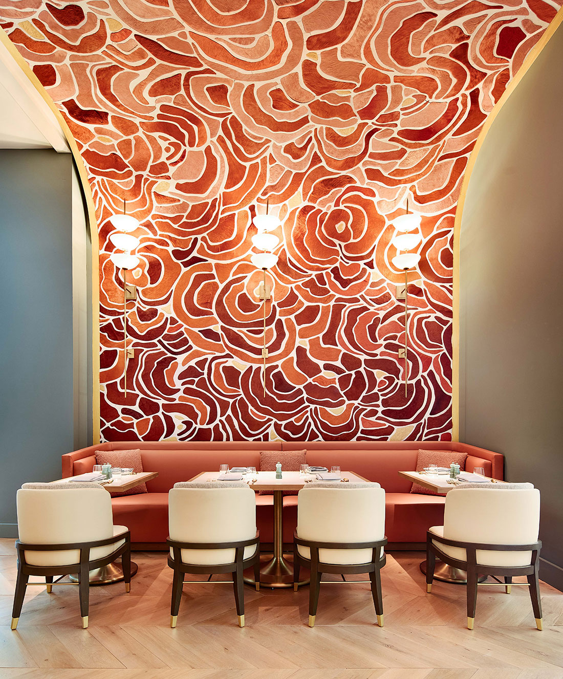 A stylish restaurant features a bold, floral-patterned ceiling in warm tones. A plush, curved booth with red upholstery is set against the wall, accompanied by four white chairs around small tables. Warm lighting adds to the elegant ambiance.