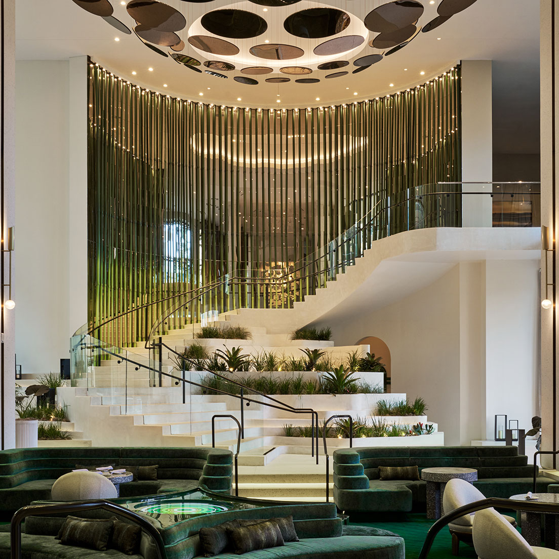 A modern, luxurious lobby with a grand staircase featuring lush greenery. The staircase has glass railings and leads to a mezzanine adorned with vertical metal rods and circular lighting fixtures on the ceiling. Sofa seating area and lush plants decorate the space.