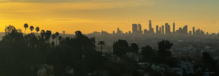 Panoramic view of a city skyline during sunset, with a golden sky casting a warm glow over the high-rise buildings. Silhouetted trees, including palm trees, are visible in the foreground, and there are residential buildings at the base of the hills.
