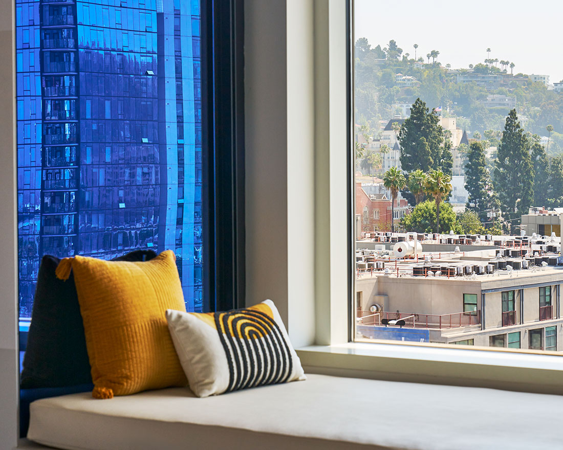 A window seat with a white cushioned bench and two pillows—one yellow and one with a black, white, and yellow spiral pattern. Outside, a modern blue building and a view of the city with trees and hills in the distance can be seen.