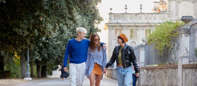 Three young adults walk outdoors on a pathway, smiling and talking. Two people have their arms around the person in the center. There are trees on one side and a historic building with a fence on the other.
