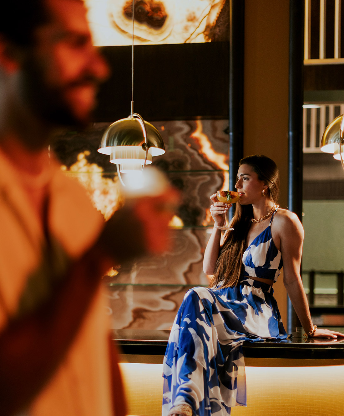 A woman in a blue and white dress sits on a bar counter, holding a drink. She looks to the side, with long hair over one shoulder. A blurred person in the foreground also holds a drink. Warm lighting fills the scene.