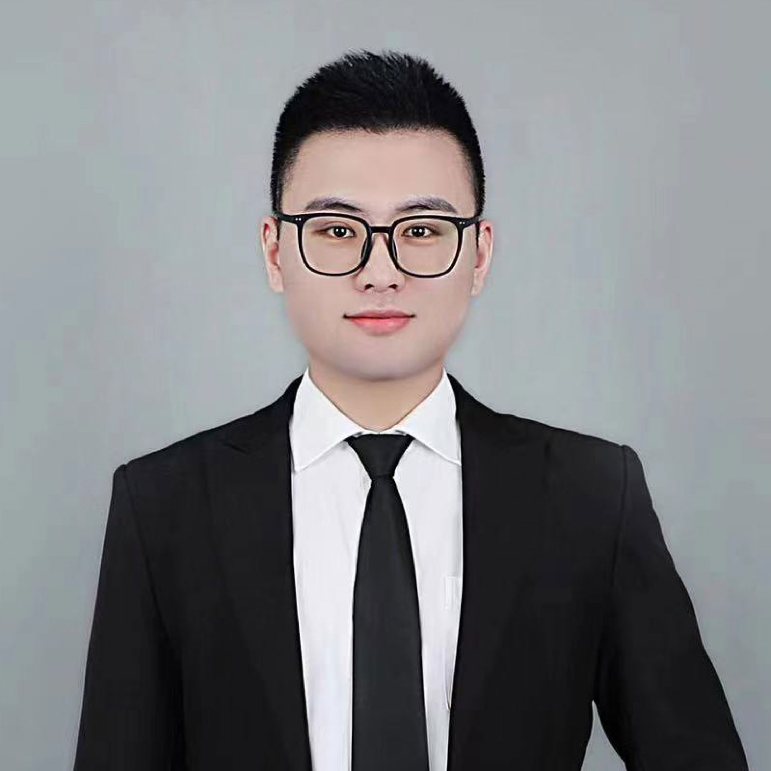 A person in a black suit, white dress shirt, and black tie stands against a gray background. They have short black hair and are wearing black-framed glasses.