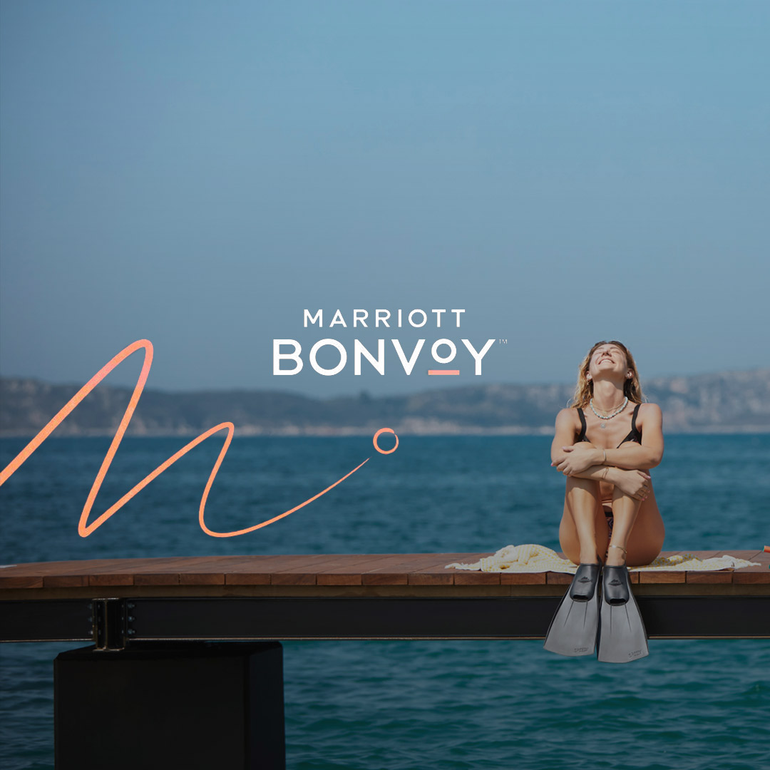 A woman sits on a dock by the sea, smiling and basking in the sun, with a large bag beside her, under a clear sky. the marriott bonvoy logo appears in the top right corner.