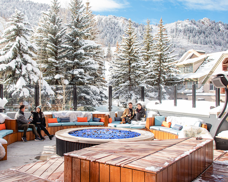 A group of people relaxing by a large fire pit in an outdoor lounge area surrounded by snow-covered trees and mountains.