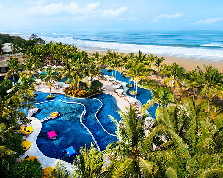 Aerial view of a tropical beach resort with a winding blue pool surrounded by palm trees, overlooking a sandy beach and the ocean.