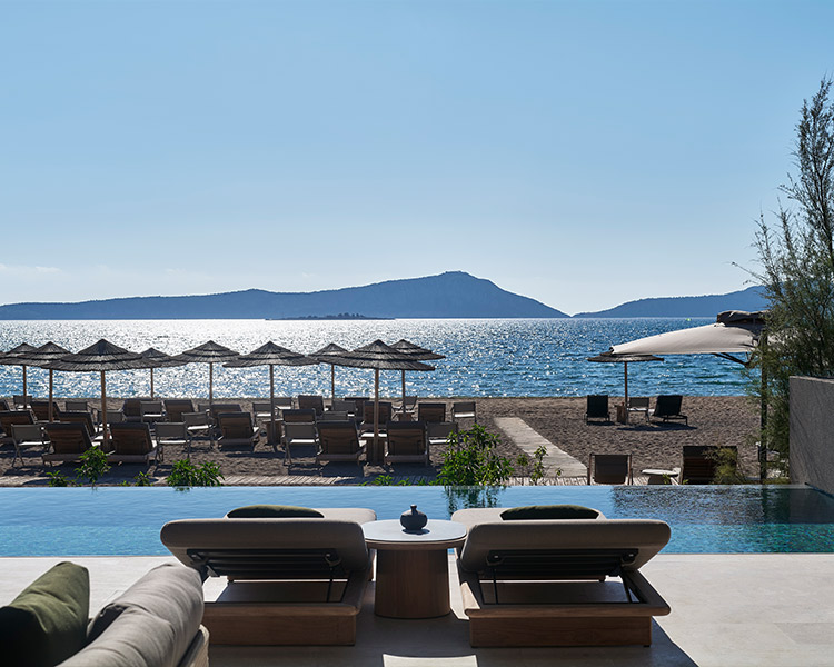 A luxury resort pool with sun loungers and umbrellas facing a tranquil sea with a mountain in the distance under a clear blue sky.