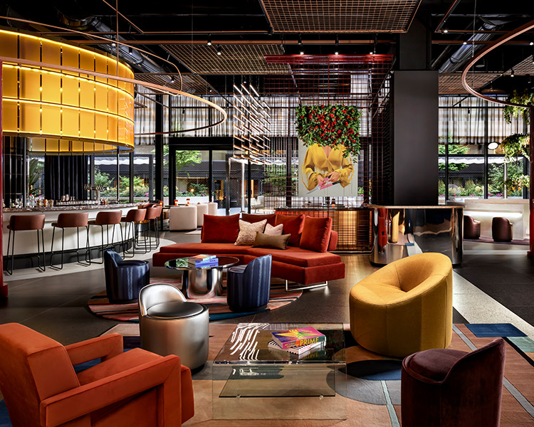 A stylish and modern lounge with eclectic seating, a central bar with a glowing yellow canopy, rich wooden accents, and large windows overlooking greenery. vibrant art pieces and plants enhance the ambiance.