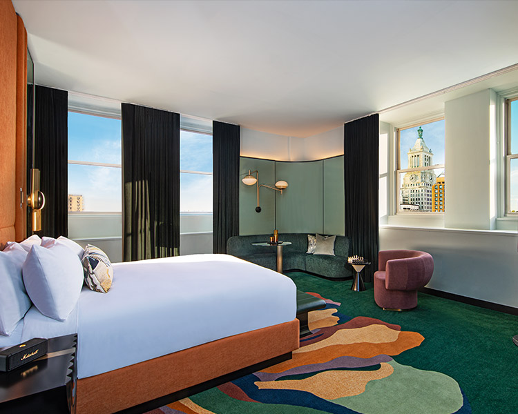 A stylish hotel room with a large bed, teal walls, and dark curtains. the room features a unique multicolored carpet, contemporary furniture, and large windows showcasing a city view.