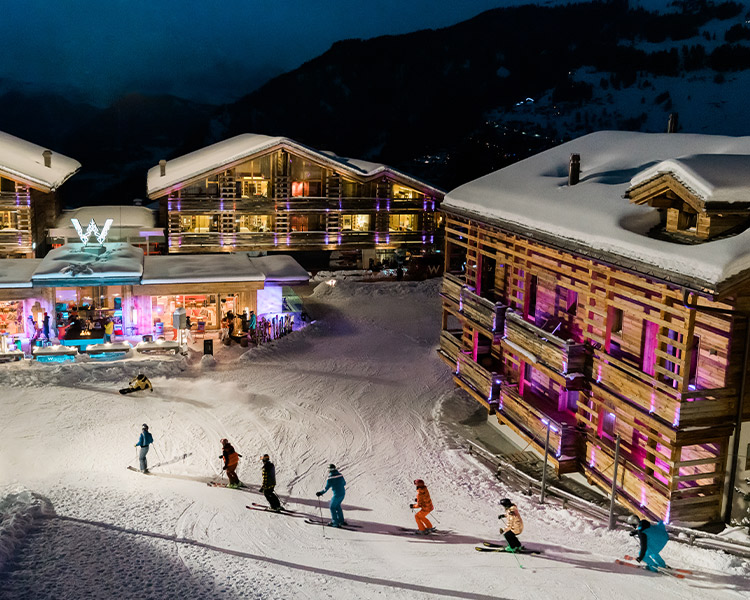 A group of skiers glides past a brightly lit, upscale lodge adorned with neon lights at a snowy mountain resort during twilight.