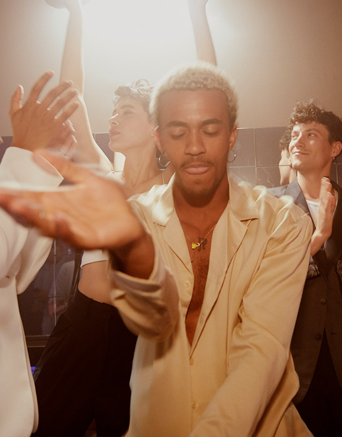 A group of young adults dancing joyfully in a dimly lit room. the focus is on a man in the foreground with extended arms, wearing a light beige suit and smiling subtly.
