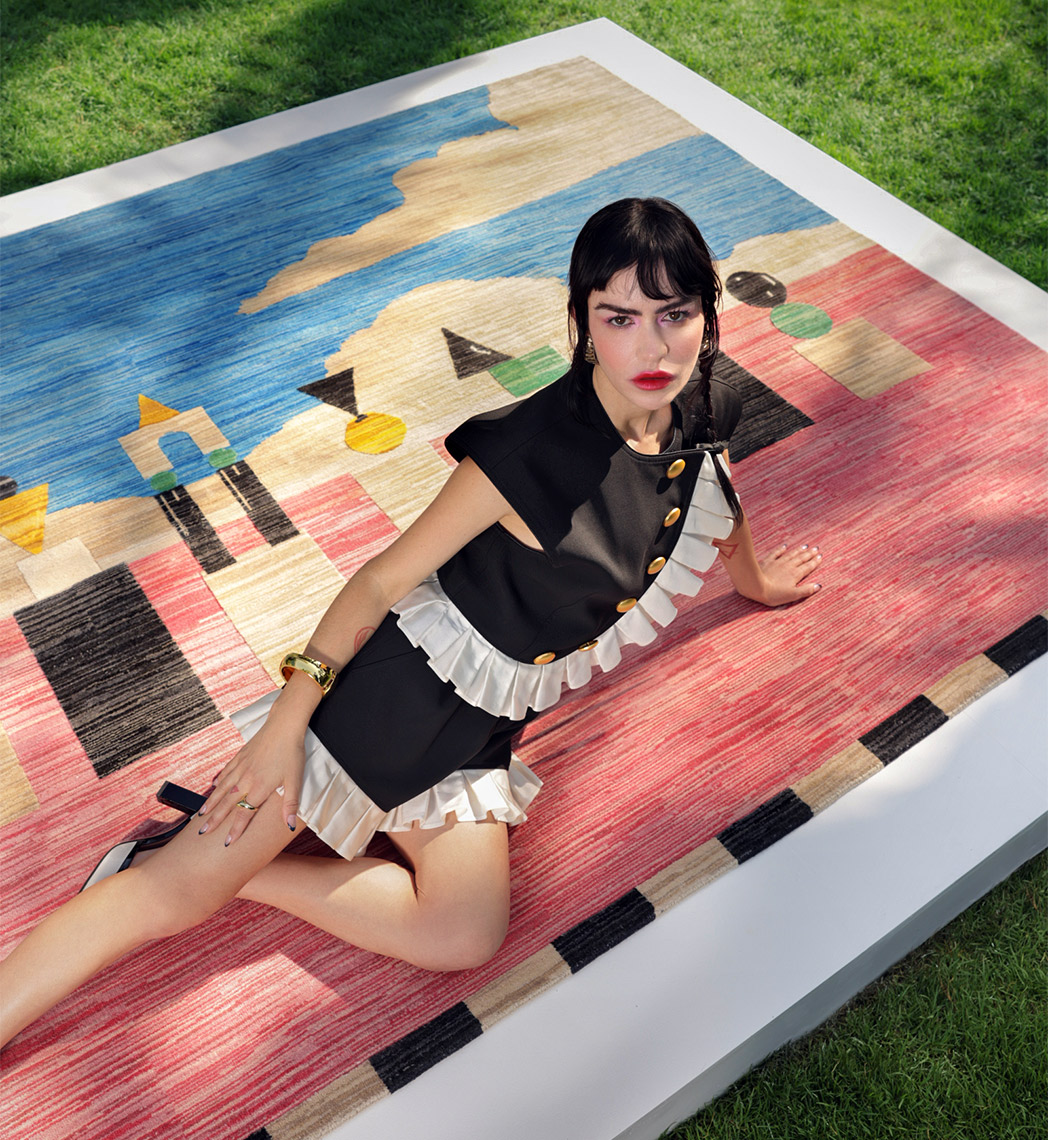 A woman with dark hair and bangs sits on a large, colorful abstract rug placed on grass, wearing a stylish black and cream dress, accessorized with bold makeup and a gold bracelet.
