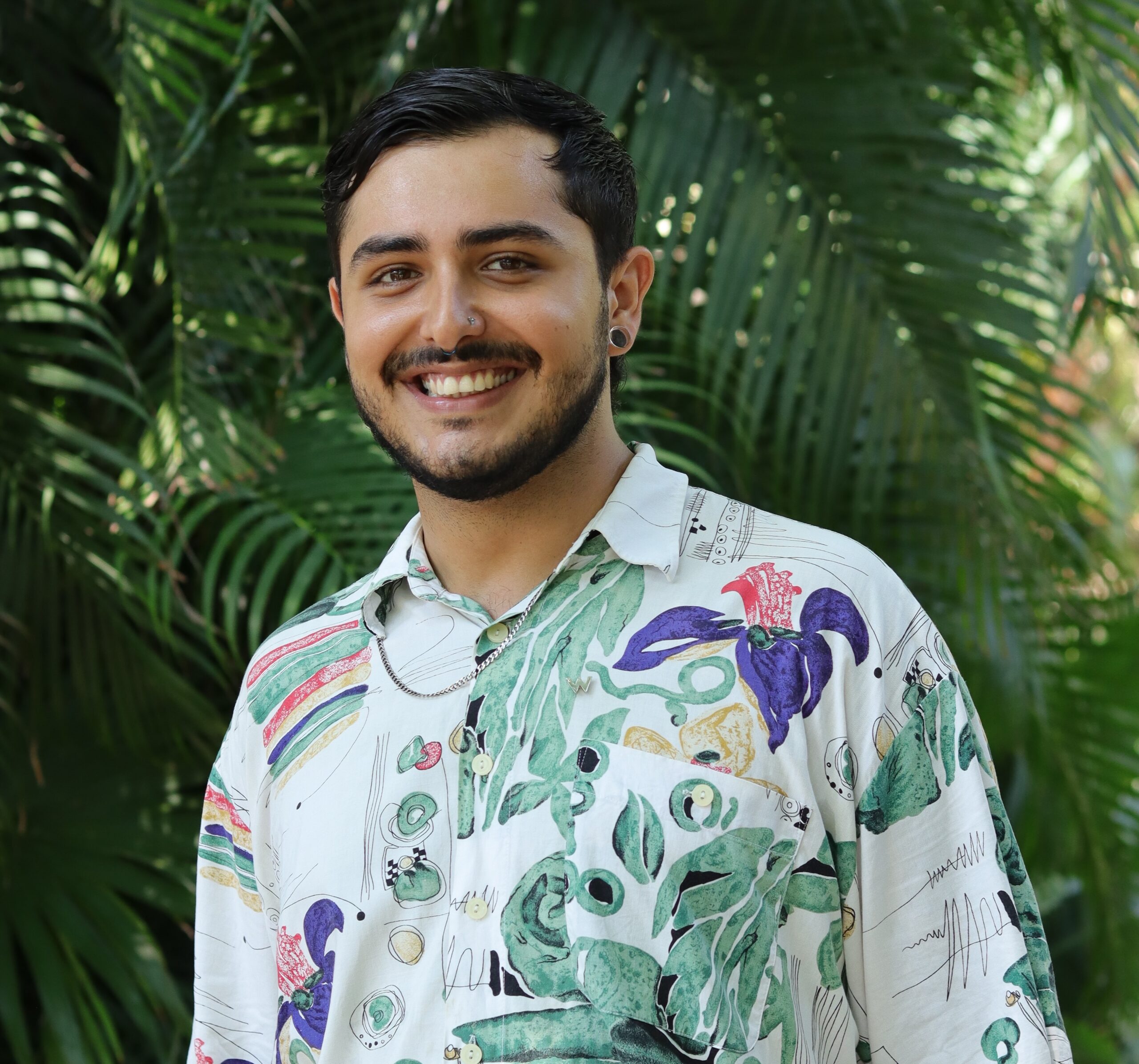 A person with short dark hair and a beard smiles at the camera. They are wearing a colorful, patterned shirt with various illustrations of plants and shapes. The background is filled with lush green foliage.