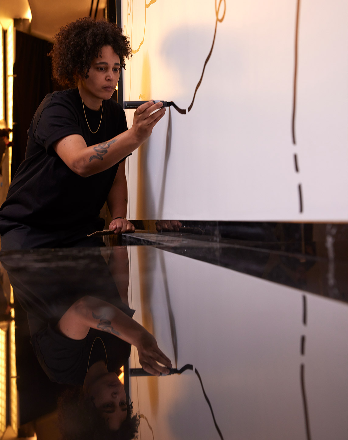 A person with curly hair is marking lines with a pen on large reflective sheets laid on a table, with their focused reflection visible on the glossy surface.