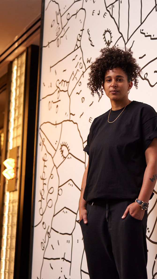 A person with curly hair, standing confidently in a black outfit, next to a large, white wall featuring an abstract black line drawing. a golden light fixture glows softly in the background.