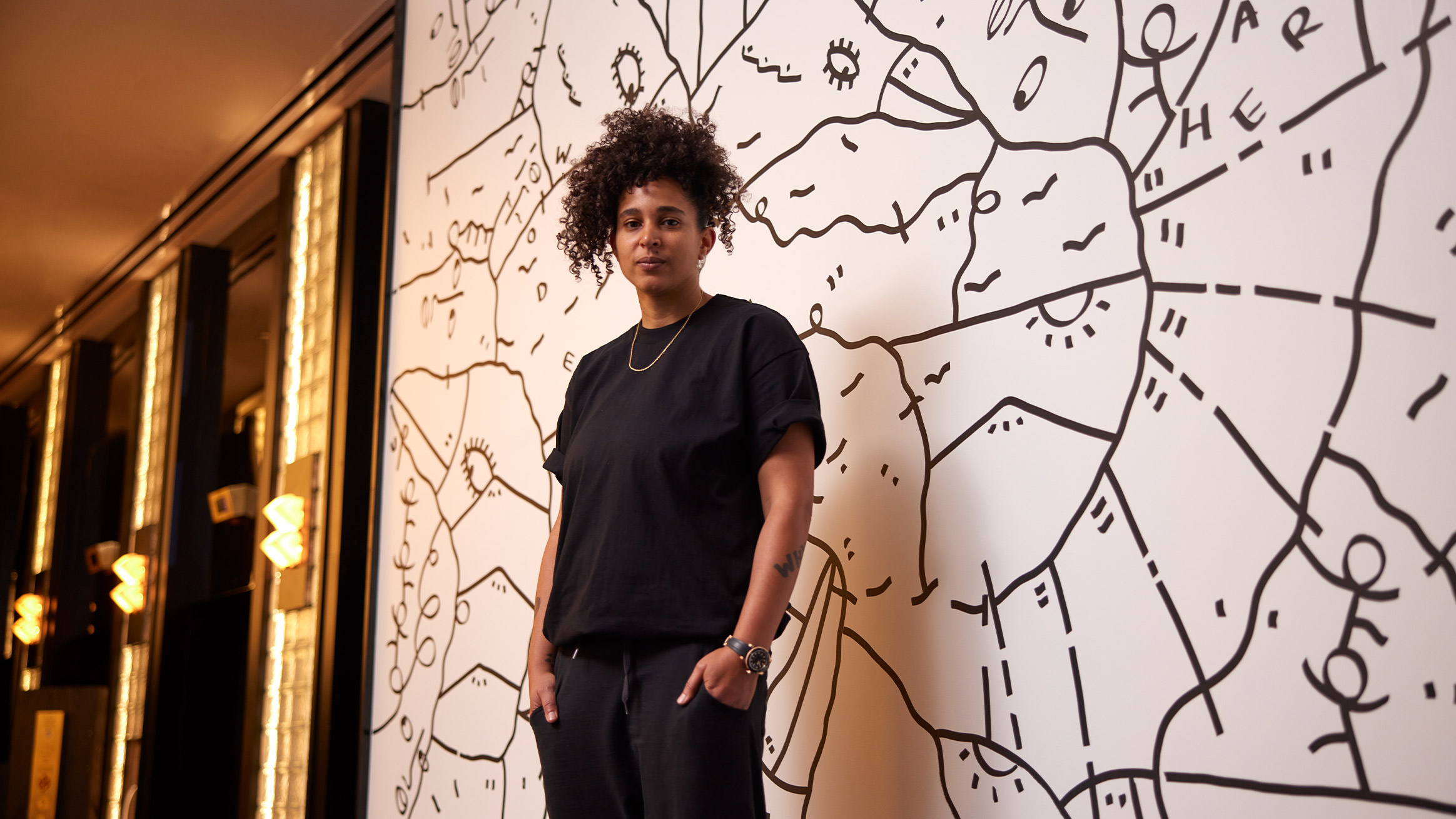 A woman with curly hair stands confidently in front of a wall with a black and white abstract doodle. she wears a black t-shirt and jeans, looking directly at the camera.