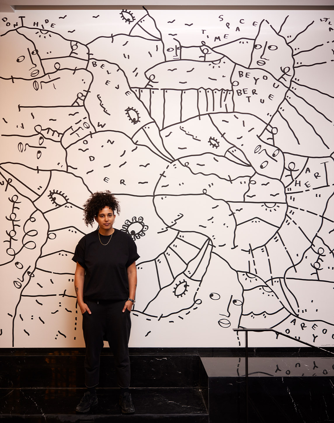 A young person with curly hair standing in front of a large, black and white abstract mural filled with various shapes and doodles. the individual wears a black outfit and poses confidently.