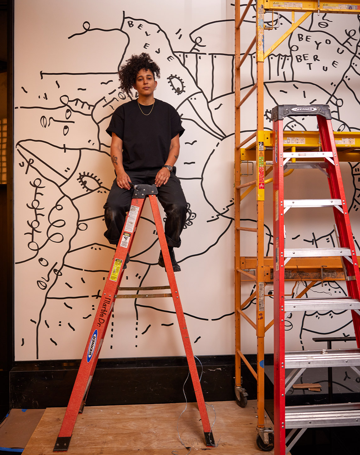 An artist sits on a red ladder in front of a mural featuring abstract black line drawings on a white wall, flanked by another ladder and painting tools.