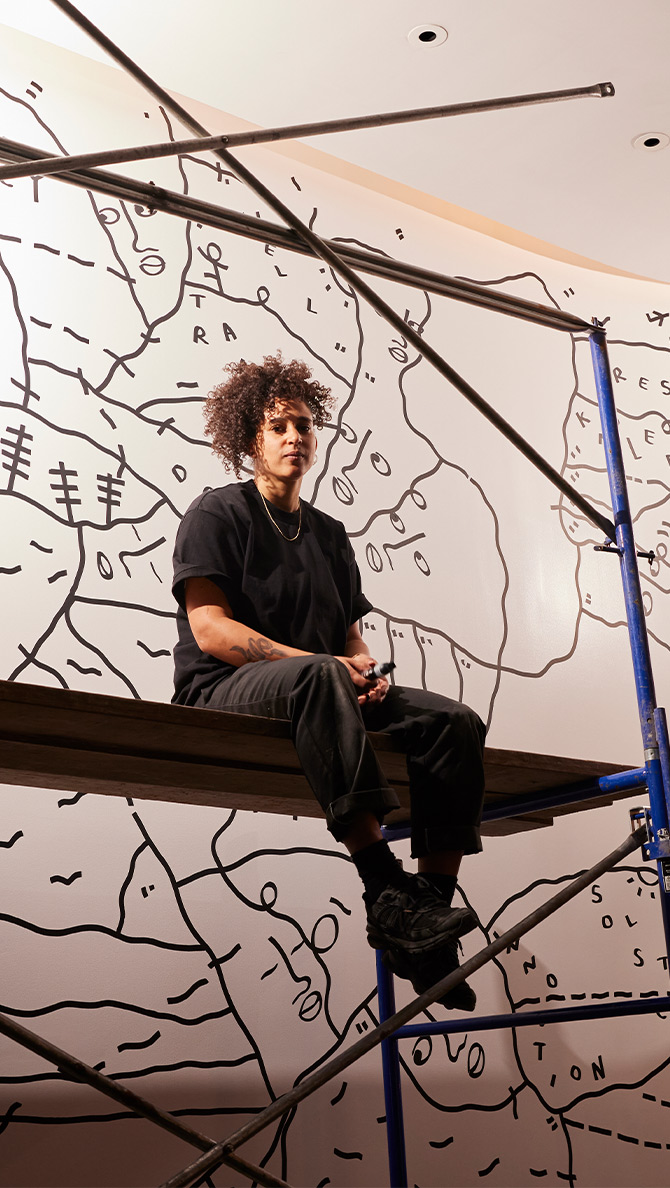 A person with curly hair and glasses, wearing a black outfit, sits on scaffolding against a wall adorned with abstract black line drawings on a white background. The scaffolding consists of metal bars and a wooden plank.